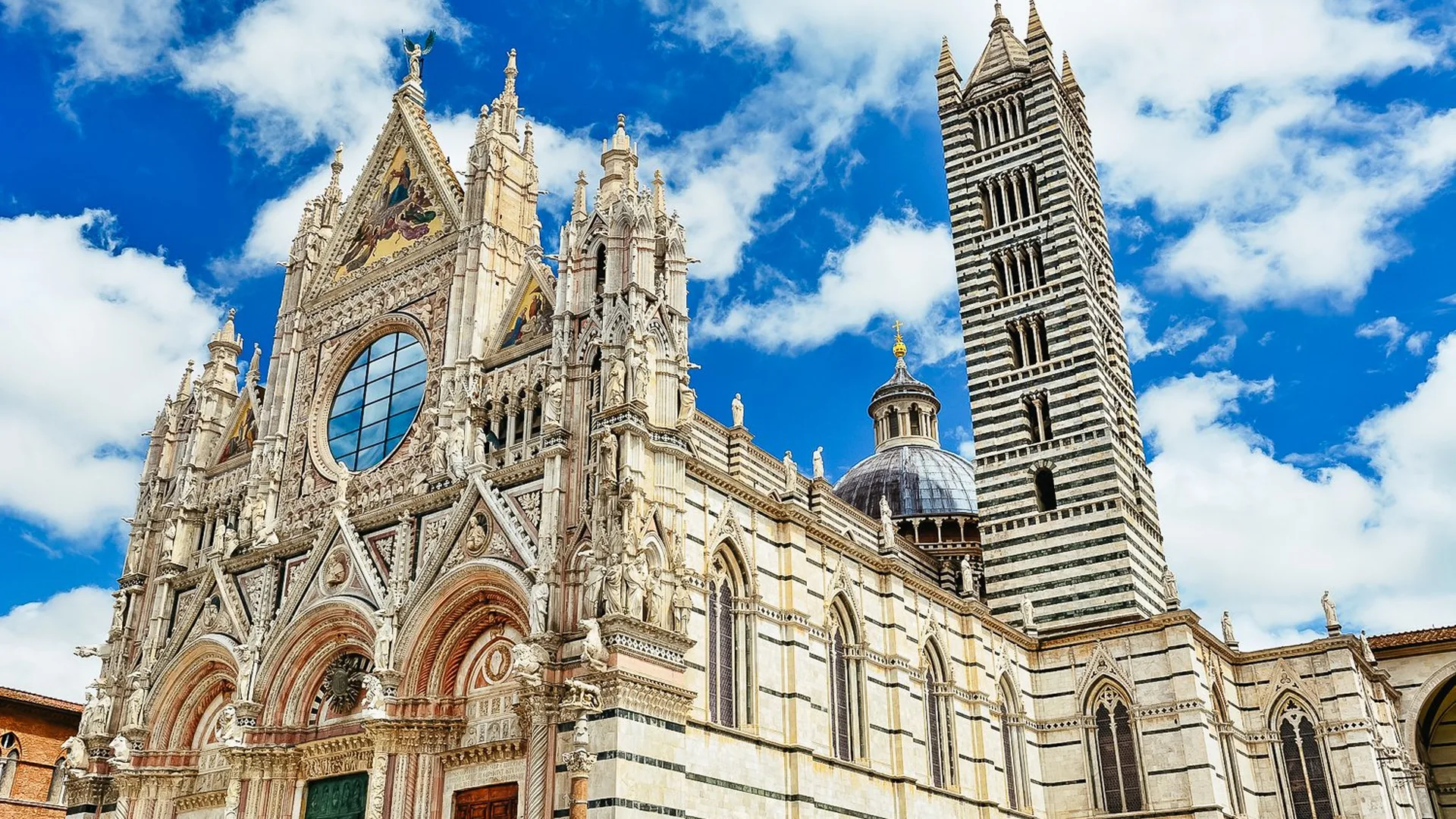 Siena Cathedral's marble facade and its black-and-white striped bell tower under a blue sky with clouds, from the OPA SI PASS listing