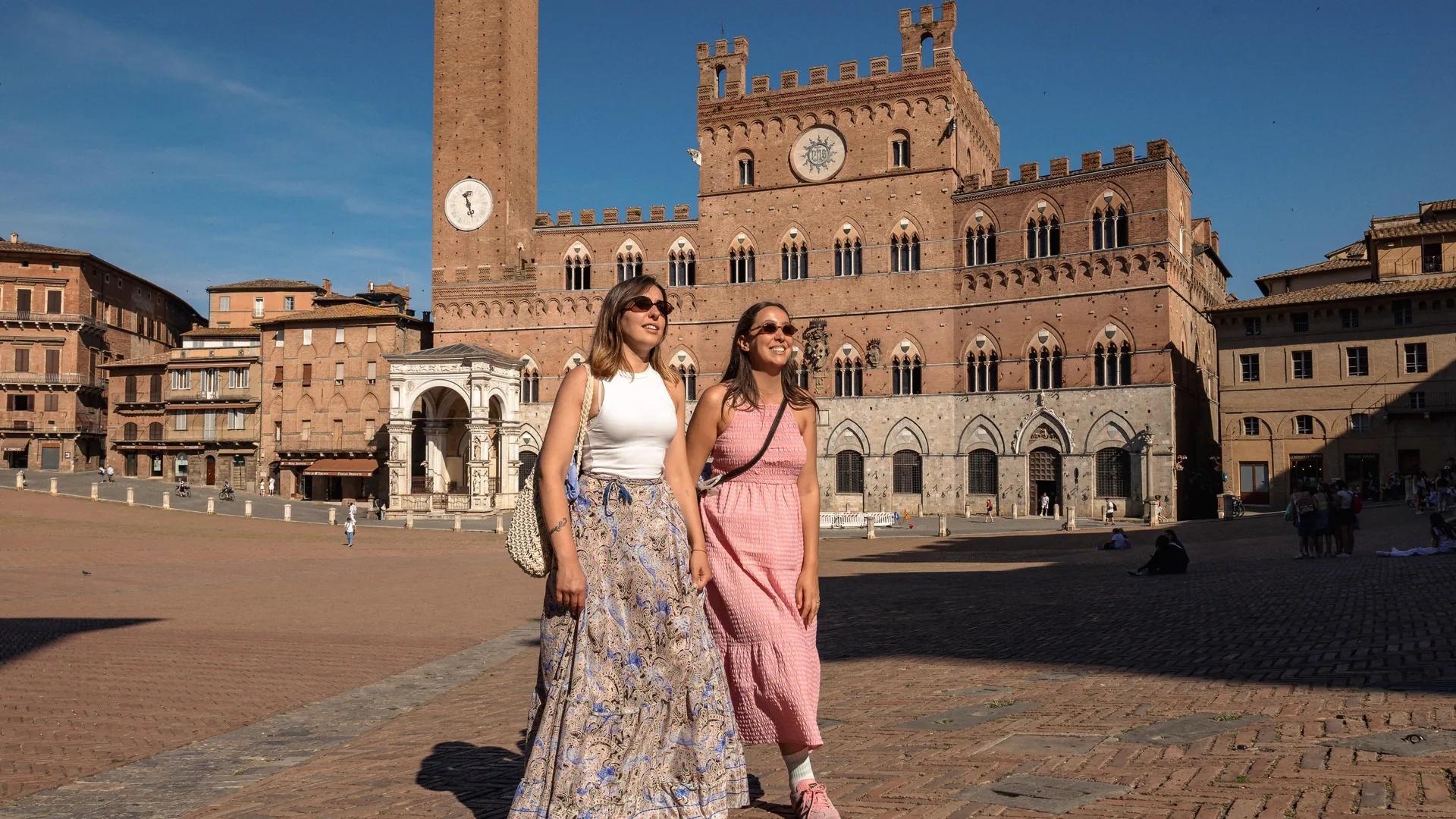 Two visitors walking across Piazza del Campo in Siena with the Palazzo Pubblico and the Torre del Mangia behind, from the day trip listing